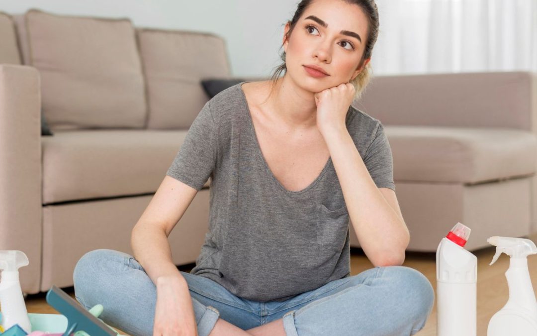 Mujer sentada en el piso de su sala, rodeada de productos de limpieza, mirando pensativa mientras considera cómo eliminar malos olores del hogar.